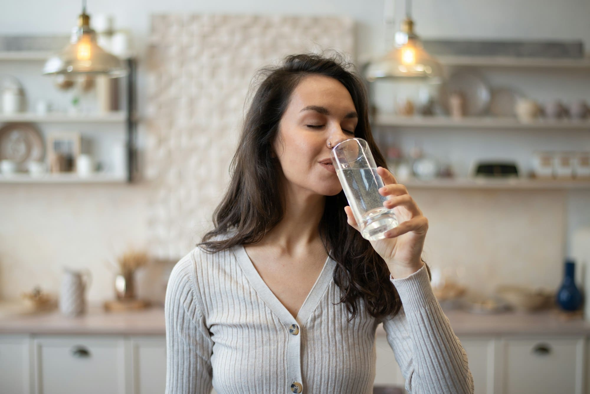 Young caucasian woman drinking spring fresh water at home, standing in kitchen, hydrating her body